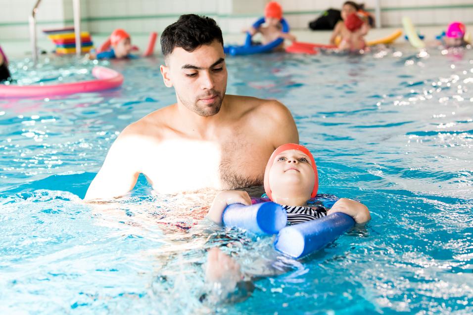 Mann unterstützt ein Kind mit Schwimmhilfen im Wasser eines Schwimmbads, während andere Schwimmer im Hintergrund sind.