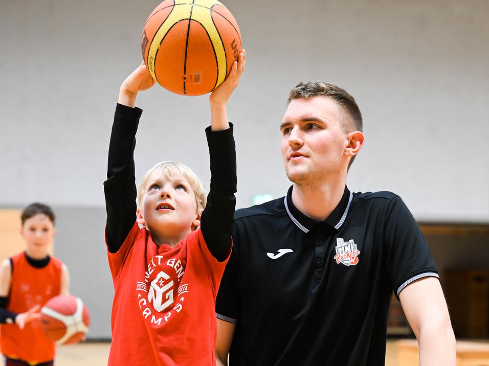 Ein junger Basketballspieler hebt den Ball über seinen Kopf, während ein Trainer neben ihm kniet und zuschaut.