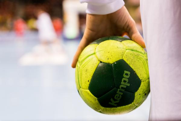 Nahaufnahme einer Hand, die einen grünen Handball mit gelben Elementen hält, im Hintergrund Spieler auf dem Spielfeld.