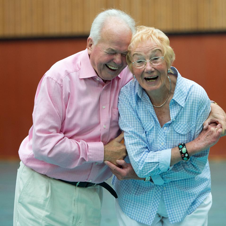 Lächelndes älteres Paar umarmt sich in einer Turnhalle, umgeben von Holzwand und sportlichem Ambiente.